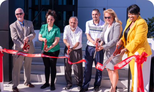 Image of six people cutting a grand opening ribbon in front of a building.